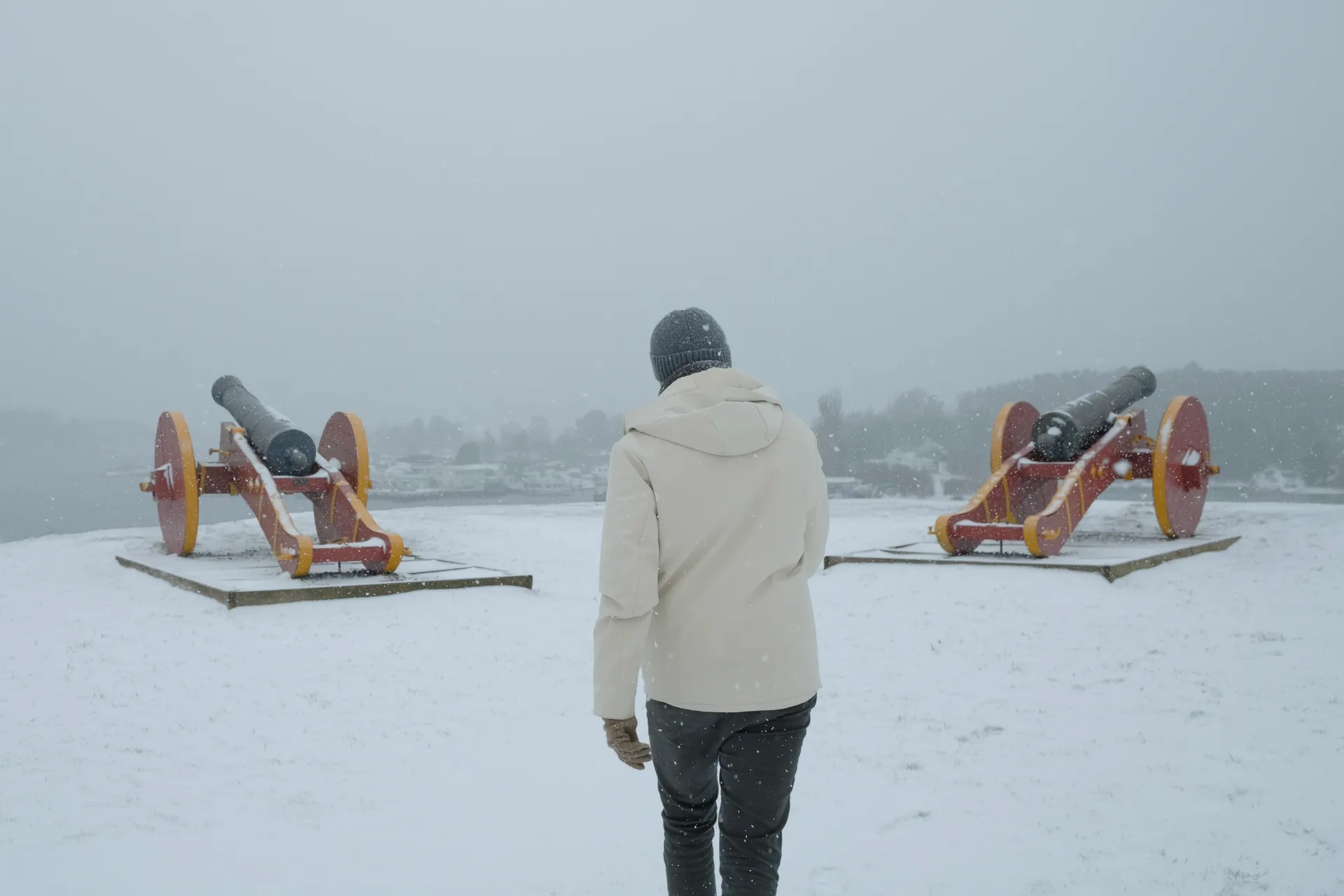 A person in a white coat walking between two old cannons in the snow.
